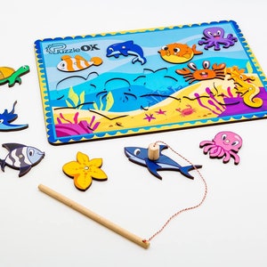 Può includere: Un gioco di pesca in legno a tema marino con un tabellone blu e giallo e creature marine colorate. Il gioco include una canna da pesca con una corda e una calamita. Le creature marine includono un delfino, un pesce, un granchio, un cavalluccio marino, un polpo, una razza e una tartaruga.