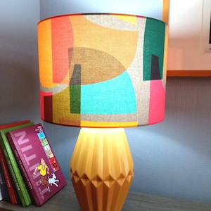 May include: A decorative table lamp featuring a lampshade with a colourful geometric pattern in shades of orange, green, pink, and blue. The mustard yellow base has a faceted design. Books are visible on a shelf.