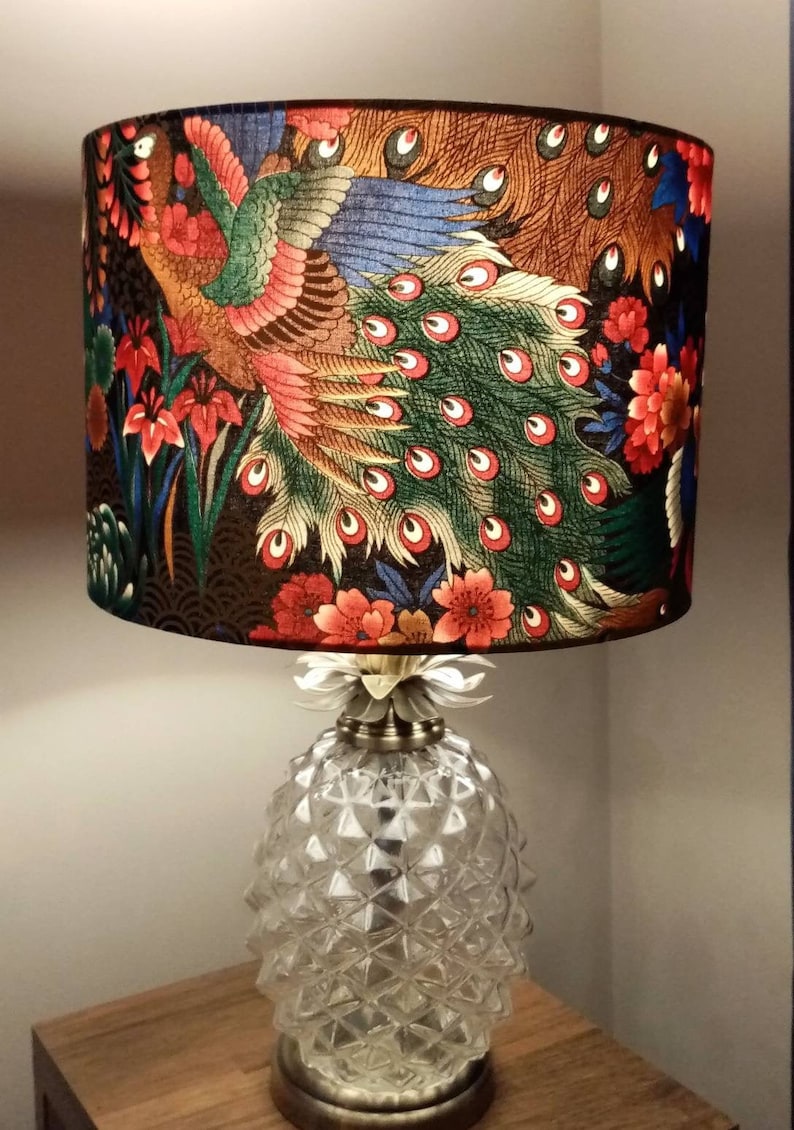 May include: A table lamp with a pineapple-shaped base and a lampshade featuring a colorful peacock design. The lampshade is made of fabric and has a black background with red, green, blue, and gold accents.