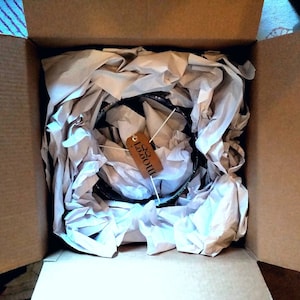 May include: A brown cardboard box with a white paper packing material inside. The packing material is crumpled and has a brown tag with the word "Moooi" written on it.