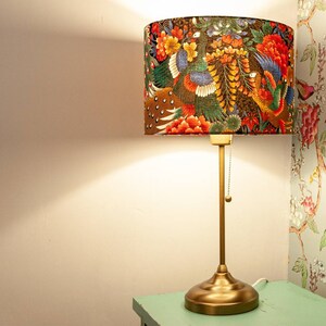 May include: A gold table lamp with a fabric lampshade featuring a colorful floral and bird design. The lampshade has a brown background with red, orange, yellow, green, and blue flowers and birds.