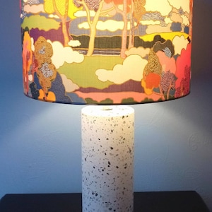 Pode incluir: Uma base de l&acirc;mpada branca com um padr&atilde;o salpicado e um abajur colorido com um design de paisagem que apresenta &aacute;rvores e nuvens.