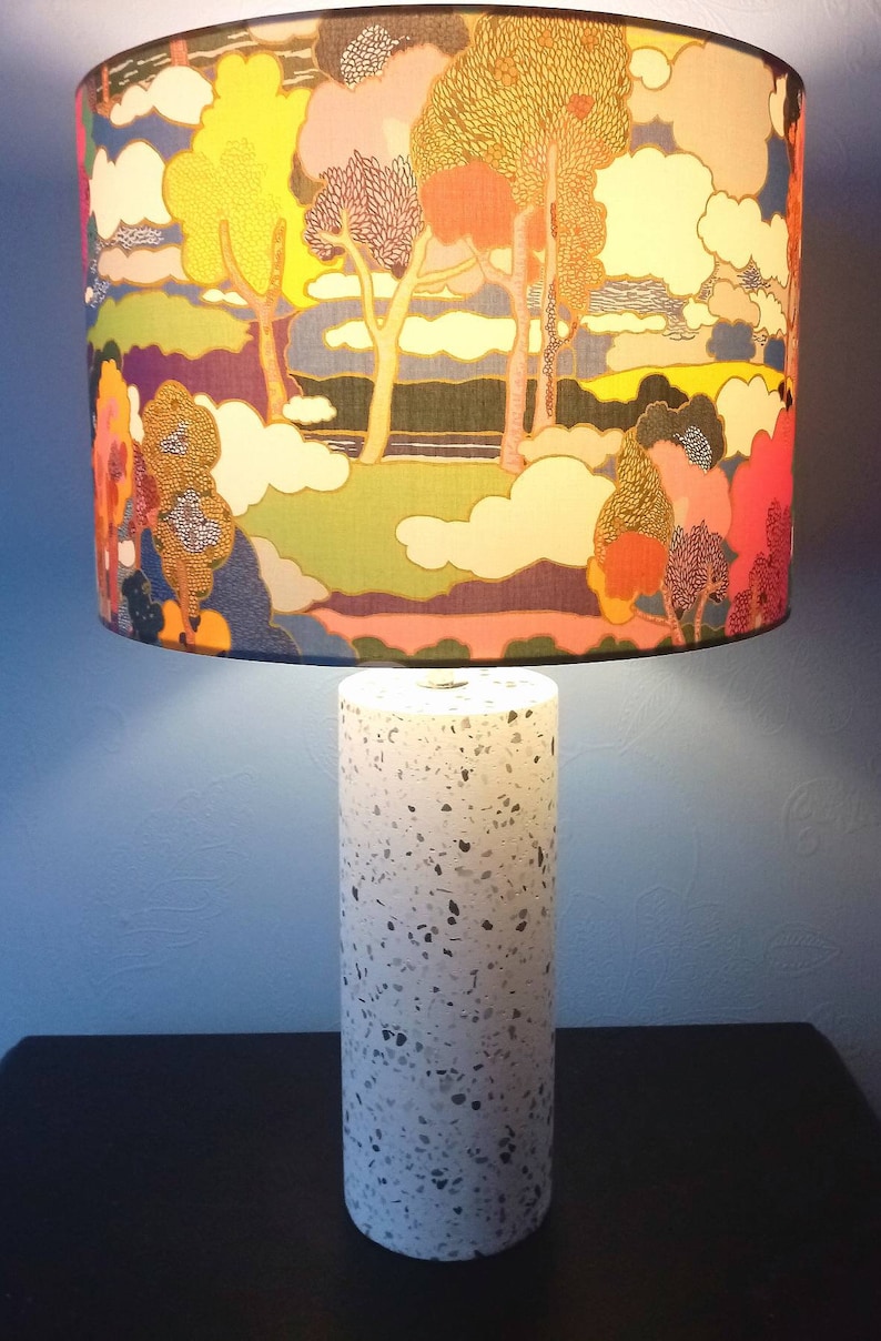Pode incluir: Uma base de l&acirc;mpada branca com um padr&atilde;o salpicado e um abajur colorido com um design de paisagem que apresenta &aacute;rvores e nuvens.