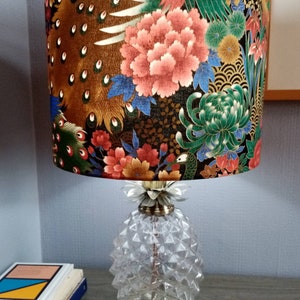 May include: A table lamp with a pineapple-shaped base and a fabric lampshade featuring a floral and peacock feather design in shades of gold, green, blue, red, and pink.