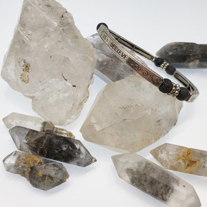 May include: A silver bracelet with black lava beads and the word "BELIEVE" engraved on the band. The bracelet is displayed on a white surface with several clear quartz crystals.