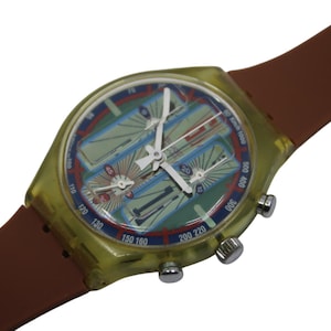 Pode incluir: Um relógio de plástico transparente amarelo com uma pulseira de couro marrom. O mostrador do relógio tem um design multicolorido com três mostradores de cronógrafo.