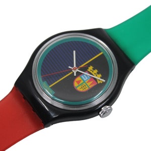 May include: A black and green wristwatch with a red and green strap. The watch face has a blue and yellow design with a crest in the center.