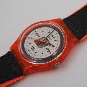 Puede incluir: Un reloj de pulsera naranja brillante con una esfera blanca y detalles en negro. El reloj tiene una correa de tela naranja con secciones de cuero negro. La esfera muestra la hora con números y agujas negras. Una etiqueta está adjunta.