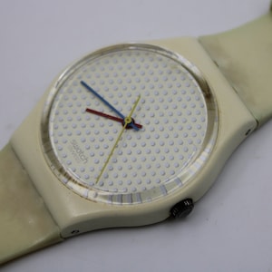 Könnte beinhalten: Eine weiße Swatch-Uhr mit weißem Armband und weißem Zifferblatt mit einem Muster aus kleinen weißen Punkten. Die Uhr hat einen blauen, roten und gelben Sekundenzeiger.