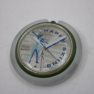May include: A silver and green Swatch watch with a white face. The face has a skier in blue and white on it. The words "Happy Skiing" are printed around the edge of the face. The words "Ga-Chamonix, St. Moritz, Kitzbuhel, Lech, Aros, Innsbruck, Courchevel, Wini" are printed around the skier.