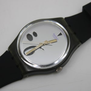 May include: A black and silver Swatch watch with a white face and black accents. The watch has a black rubber band.