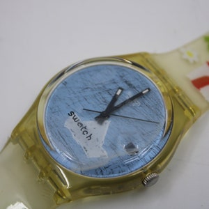 May include: A yellow plastic Swatch watch with a blue face and black hands. The watch face has a white paper-like design with the word "swatch" written on it. The watch band has a white and red house design with green trees and white flowers.