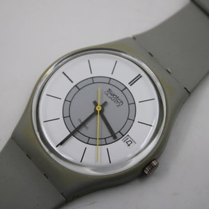 May include: A grey Swatch watch with a white face and a grey strap. The watch face has a smaller grey circle in the centre with the words "Swatch Quartz" printed on it. The date window is at the 4 o'clock position and shows the number 14.