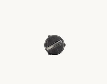 May include: A circular black metal screw cap featuring a flat top surface with a prominent horizontal slot across the centre. The cap has a smooth, dark finish and small, evenly spaced protrusions along the outer edge of its circular perimeter.