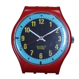 May include: A red plastic wristwatch with a black face and blue outer ring. The watch face has yellow numbers and the text "Swatch Swiss" printed on it.
