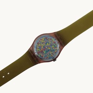 Op de afbeelding: Een vintage polshorloge met een transparante kast en een kleurrijke, abstracte wijzerplaat. Het horloge heeft een lichtblauwe wijzerplaat met een patroon van gele, roze en blauwe vormen. Het heeft een roze secondewijzer en een groene band.