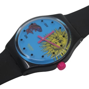 May include: A Swatch watch with a black strap and case. The watch face is light blue with a sun graphic and a fish graphic. The hands are pink. The watch has a pink crown.