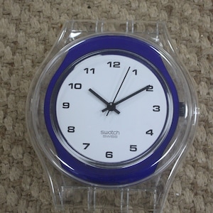 May include: A clear plastic watch with a blue face and white numbers. The watch face says "Swatch Swiss".