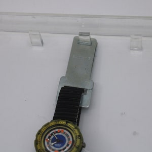 May include: A green and black wristwatch with a blue face and orange accents. The watch is on a black rubber band and is attached to a silver metal tag that says "Swatch".