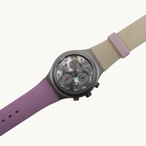 May include: A wristwatch featuring a round silver-tone case and a mother-of-pearl dial with crystal hour markers. The watch includes three sub-dials and a two-tone silicone strap that transitions from a light purple to a pale cream color.