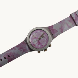 May include: A wristwatch featuring a pink and white camouflage pattern on the strap and dial. The watch has a round silver-toned case, three gold-toned side buttons, and silver-toned hands. The face includes three sub-dials and a pink background.