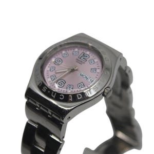 May include: A silver-toned Swatch watch with a pink face. The watch has a date display showing "MON 16". The watch band is also silver-toned. The watch face has white numerals and hands.