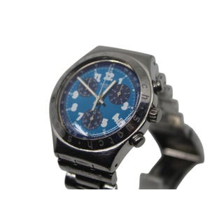 May include: A silver-toned Swatch chronograph watch with a blue dial. The watch features white numerals and hands, three sub-dials, and a date window. The watchband is made of silver-toned metal.