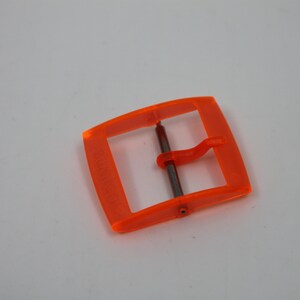 May include: Orange plastic buckle with a metal pin. The buckle is designed for a strap or belt.