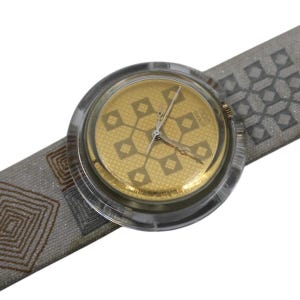 Puede incluir: Un reloj de pulsera vintage con esfera dorada y un patrón geométrico de diamantes y cuadrados. La caja es de plástico transparente y la correa de tela gris presenta un diseño geométrico repetido. Las manecillas indican las 10:10.