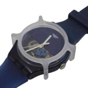 May include: A blue Swatch watch with a transparent protective case. The watch features a dark blue face with white hands and a blue strap. The watch case is a light gray color.
