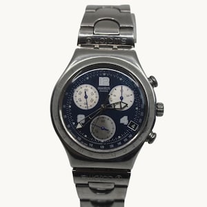 May include: A silver-toned Swatch chronograph watch with a dark blue dial. The watch features three sub-dials, white numerals, and a date display at the 4 o'clock position. The watch band is also silver-toned.