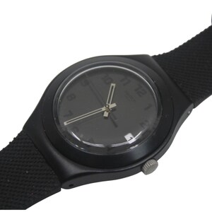 May include: Black Swatch watch with a black dial and hands. The watch has a black strap and a silver crown. The watch face displays the numbers 1 through 12. The watch is a classic and minimalist design.