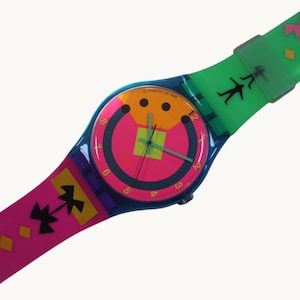 May include: A colorful Swatch watch with a blue case and a vibrant pink, orange, and green face. The watch band is pink and green with geometric and silhouette designs. The watch face has the text "SWATCH AG 1983".