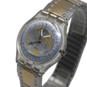 May include: A clear Swatch watch with a silver and gold-tone metal band. The watch face features a silver and gold design with French text. The watch hands are gold-tone.