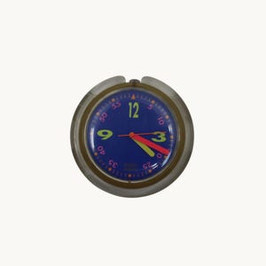May include: A round wall clock with a translucent outer frame. The blue face features large lime green numerals for 3, 9, and 12, with pink minute markers in increments of five. It has orange hour and minute hands and a thin orange second hand.