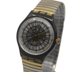 May include: A Swatch Swiss watch with a black case and a silver and gold-tone band. The watch face has Roman numerals and a skeleton design, with the brand name visible. The watch has a gold-tone crown.