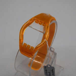 Könnte beinhalten: Orangefarbenes Armband aus Kunststoff mit einem transparenten Zifferblatt aus Kunststoff. Das Armband befindet sich auf einem transparenten Kunststoffständer. Das Etikett auf dem Ständer lautet "Jewelry by Bell 10144 HH 107".