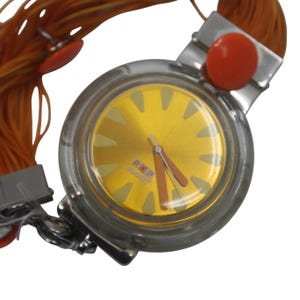 Puede incluir: Un reloj Swatch POP vintage con una caja redonda y transparente y una esfera dorada. El reloj presenta agujas e indicadores de hora naranjas, con la palabra "POP" impresa en la esfera. La correa del reloj está hecha de cordones naranjas y tiene detalles naranjas.