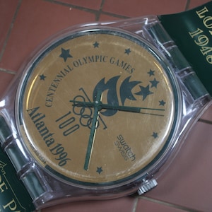 May include: A clear plastic Swatch watch with a gold face. The face features the Olympic rings, a green flame, and the text "Centennial Olympic Games Atlanta 1996 100". The watch band is green with the text "London 1948" and "Olympiade Paris" in gold.