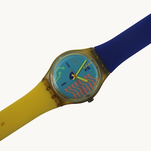 May include: A colorful wristwatch with a blue face featuring orange and green designs. The watch has a yellow and blue strap. The watch face has the numbers 3, 6, 9, and 12. The watch is a retro style.