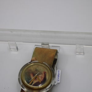 May include: A gold-toned watch with a clear plastic case and a printed strap. The watch face features a painting of a woman in a dress. The strap is a light brown and green colour with a faded floral pattern. The watch has a white tag attached to the strap that says "WATERPROOF".