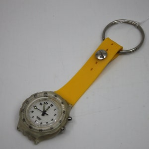 May include: A clear plastic watch with a white face and black hands. The watch is attached to a yellow rubber keychain with a metal ring.