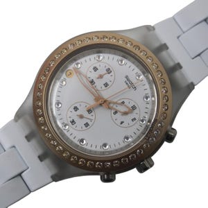 May include: A white Swatch Swiss watch with a gold-colored bezel encrusted with small, clear stones. The watch face is white with gold-colored hands and three sub-dials. The watch band is white.