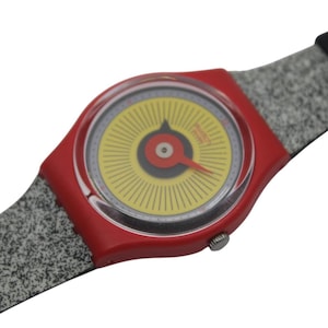 Puede incluir: Un reloj Swatch rojo con esfera amarilla y negra y aguja roja. El reloj tiene una correa moteada gris y blanca. La esfera del reloj tiene un diseño circular con líneas radiales.