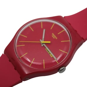 Può includere: Un orologio Swatch rosso con cinturino rosso abbinato. Il quadrante dell'orologio è rosso con indici e lancette gialle. L'orologio ha un display della data e il logo Swatch.