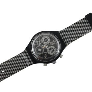 Peut inclure: Montre chronographe noire et argentée avec un bracelet à carreaux noirs et blancs. Le cadran de la montre est noir avec des détails argentés et trois sous-cadrans. Le bracelet présente un motif répété de carrés noirs et blancs.