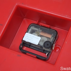 May include: Close-up of a clock mechanism with a black plastic casing and a clear face. The mechanism is labeled "USE 1.5V Carbon Zinc Battery Only NO Alkaline or Rechargeable batteries".