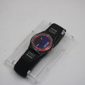 May include: A black Swatch watch with a red and blue face. The watch has a black strap with the word "Swatch" printed on it.
