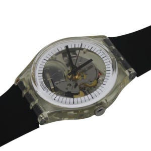 Può includere: Un orologio Swatch in plastica trasparente con un cinturino in gomma nera. Il quadrante dell'orologio è bianco con lancette nere e una finestra trasparente che mostra il funzionamento interno dell'orologio.