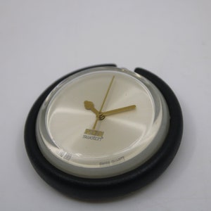 Op de afbeelding: Een ronde, zwart-witte Swatch-horloge met gouden wijzers en een witte wijzerplaat. Het horloge heeft een transparante plastic kap over de wijzerplaat.